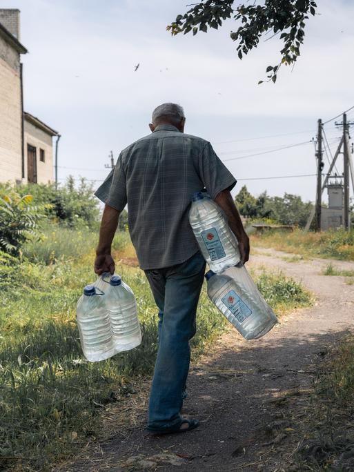 Zu sehen ist ein Mann, der große Wasserflaschen in den Händen und unter den Armen trägt. Er läuft eine unbefestigte Straße entlang, die zwischen Strommasten und einem Gebäude verläuft.