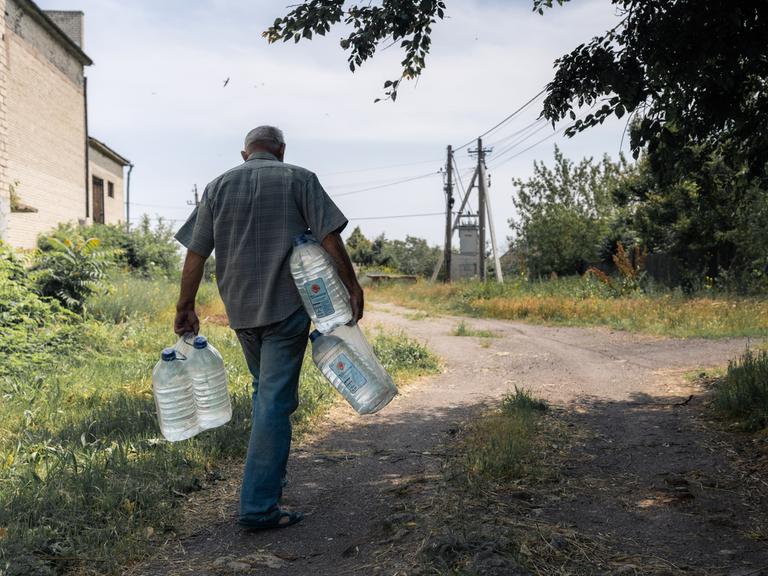 Zu sehen ist ein Mann, der große Wasserflaschen in den Händen und unter den Armen trägt. Er läuft eine unbefestigte Straße entlang, die zwischen Strommasten und einem Gebäude verläuft.