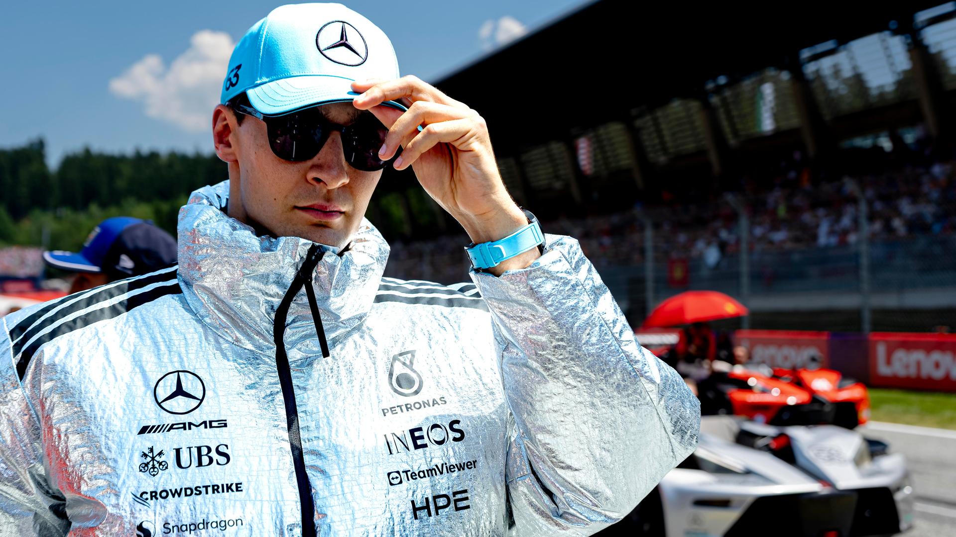 Russel steht in einer silberfarbenen Jacke und mit einer hellblauen Kappe an der Rennstrecke und blickt durch die Sonnenbrille in die Kamera.