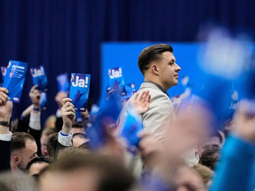 Jean-Pascal Hohm ist im Profil zu sehen, wie er zwischen anderen Parteimitgliedern steht, die Karten mit der Aufschrift "JA!" in die Luft strecken. Jean-Pascal Hohm ist im Profil zu sehen, wie er zwischen anderen Parteimitgliedern steht, die Karten mit der Aufschrift "JA!" in die Luft strecken.