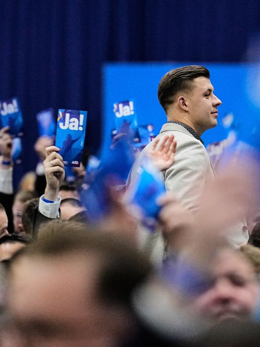 Jean-Pascal Hohm ist im Profil zu sehen, wie er zwischen anderen Parteimitgliedern steht, die Karten mit der Aufschrift "JA!" in die Luft strecken. Jean-Pascal Hohm ist im Profil zu sehen, wie er zwischen anderen Parteimitgliedern steht, die Karten mit der Aufschrift "JA!" in die Luft strecken.