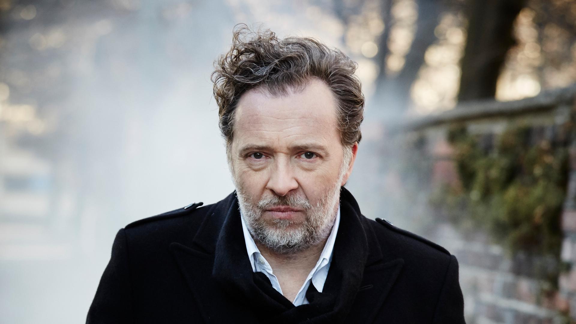 Christian Gerhaher steht im Freien. Er hat kurze, lockige Haare, trägt einen Bart und einen dunklen Mantel. Rechts ist eine Mauer zu erkennen.