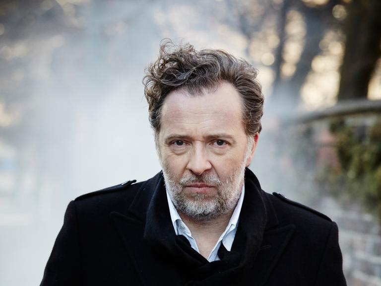 Christian Gerhaher steht im Freien. Er hat kurze, lockige Haare, trägt einen Bart und einen dunklen Mantel. Rechts ist eine Mauer zu erkennen.