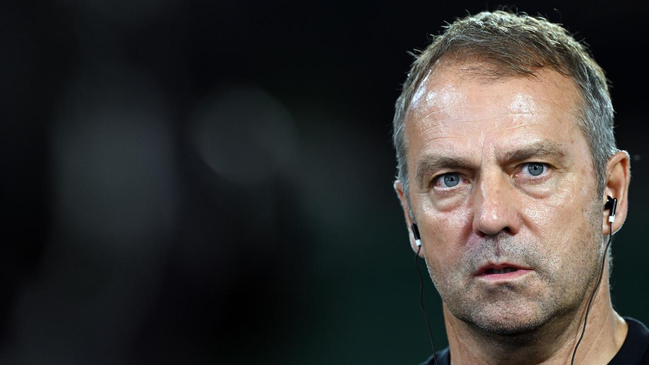 Der Trainer Hansi Flick steht in einem Stadion. Er trägt Kopfhörer im Ohr und schaut schräg nach rechts in die Kamera.