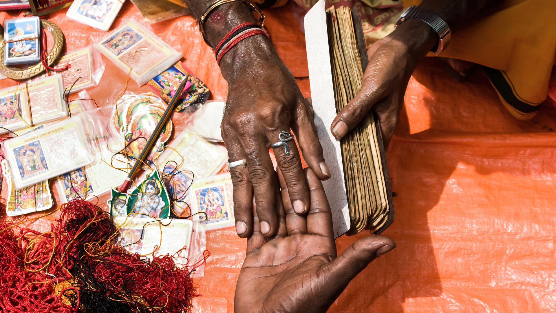 Ein Wahrsager hält die Hand seines Kunden. Daneben liegen Karten mit den Bildern hinduistischer Götter und ein Gewirr roter Schnüre.