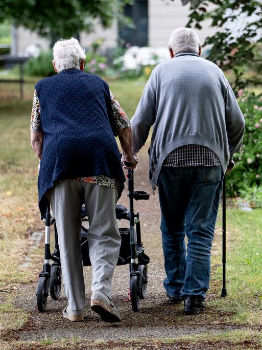 Zwei Senioren gehen mit Gehhilfen durch den Garten eines Seniorenheims. Eine Person nutzt einen Rollator, die andere einen Gehstock. Zwei Senioren gehen mit Gehhilfen durch den Garten eines Seniorenheims. Eine Person nutzt einen Rollator, die andere einen Gehstock.