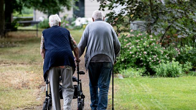 Zwei Senioren gehen mit Gehhilfen durch den Garten eines Seniorenheims. Eine Person nutzt einen Rollator, die andere einen Gehstock.
