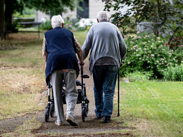 Zwei Senioren gehen mit Gehhilfen durch den Garten eines Seniorenheims. Eine Person nutzt einen Rollator, die andere einen Gehstock. Zwei Senioren gehen mit Gehhilfen durch den Garten eines Seniorenheims. Eine Person nutzt einen Rollator, die andere einen Gehstock.