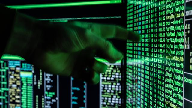 Ein Mann hält seine Hand vor ein fiktives Hacker-Programm, das auf zwei Bildschirmen eines Computers zu sehen ist.