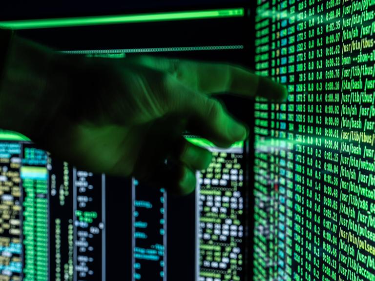 Ein Mann hält seine Hand vor ein fiktives Hacker-Programm, das auf zwei Bildschirmen eines Computers zu sehen ist.