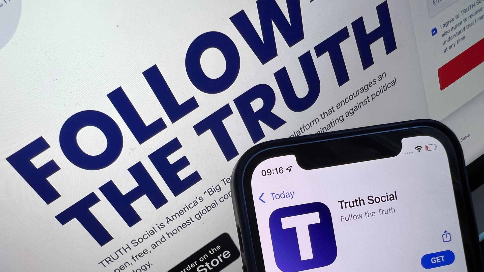 Das Bild zeigt ein Handy, auf dem die "Truth Social"-Anwendung geöffnet ist. Dahinter auf einem Bildschirm die Ankündigung des Starttermins. Das Bild zeigt ein Handy, auf dem die "Truth Social"-Anwendung geöffnet ist. Dahinter auf einem Bildschirm die Ankündigung des Starttermins.