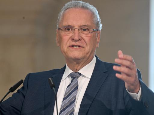 Der Bayerische Innenminister Joachim Herrmann spricht mit erhobener Hand vor zwei Mikrofonen.