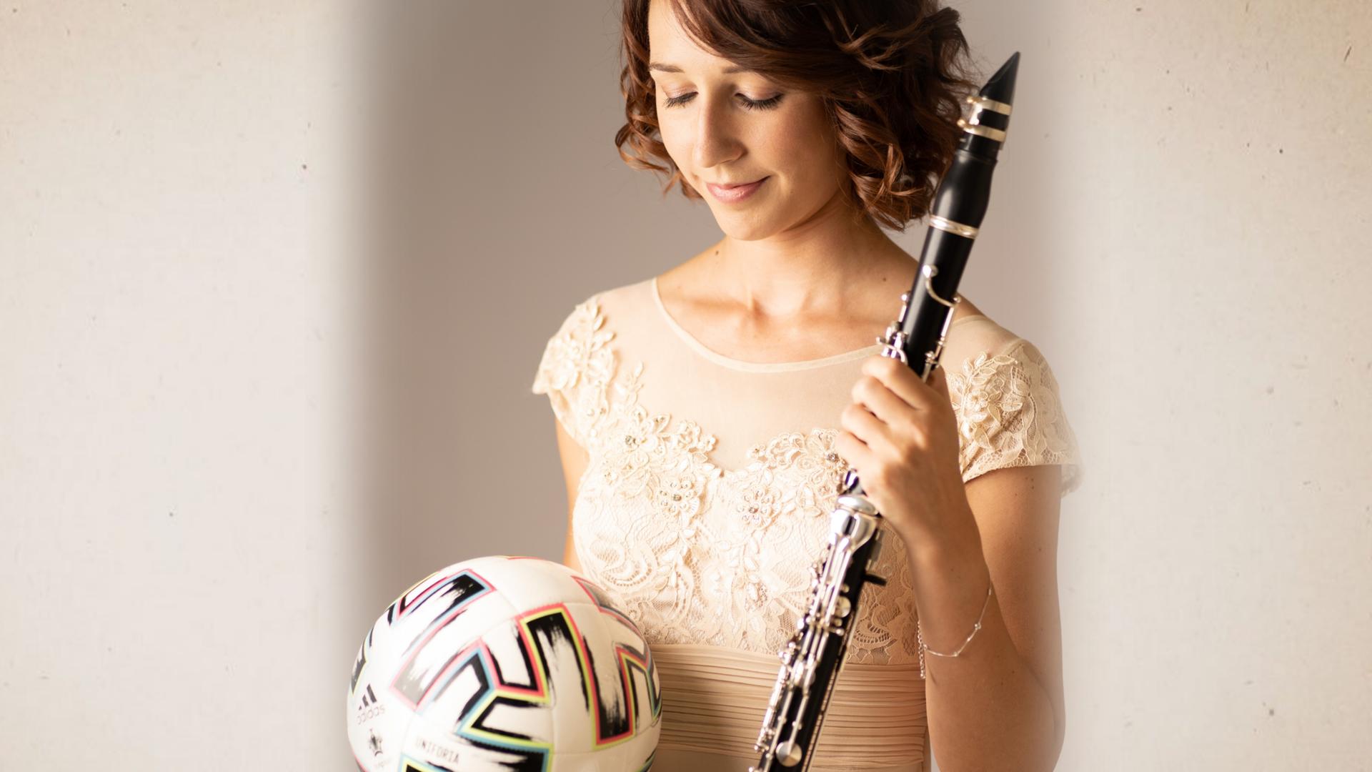 Die Klarinettistin Andrea Götsch hat in der einen Hand einen Fußball, auf den sie ihren Blick richtet, während sie in Abendrobe in der anderen Hand ihre Klarinette hält.