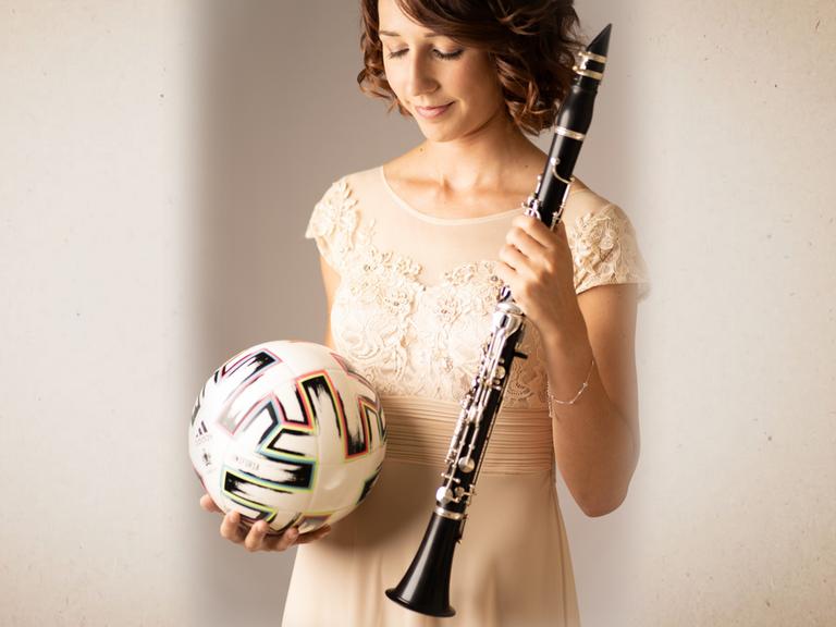 Die Klarinettistin Andrea Götsch hat in der einen Hand einen Fußball, auf den sie ihren Blick richtet, während sie in Abendrobe in der anderen Hand ihre Klarinette hält.
