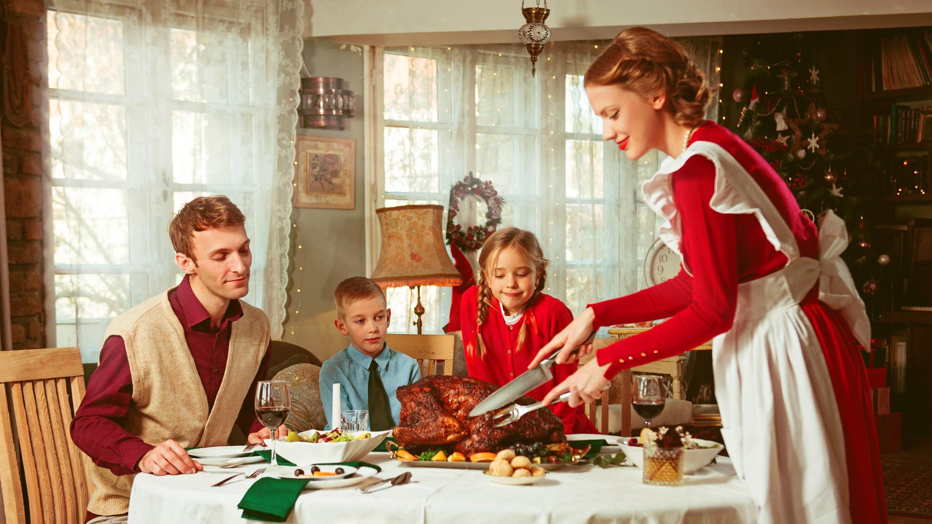 Familie bei einem Festmahl. Eine Frau im Look der 50er-Jahre schneidet einen prächtigen Braten an, auf den Mann und zwei Kinder freudig schauen. Familie bei einem Festmahl. Eine Frau im Look der 50er-Jahre schneidet einen prächtigen Braten an, auf den Mann und zwei Kinder freudig schauen.