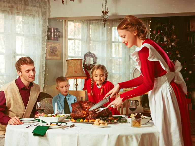 Familie bei einem Festmahl. Eine Frau im Look der 50er-Jahre schneidet einen prächtigen Braten an, auf den Mann und zwei Kinder freudig schauen.