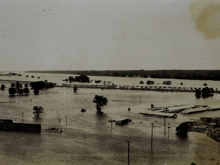 Die historische Schwarz-Weiß-Fotografie aus dem Jahr 1927 zeigt Wassermassen, aus denen vereinzelt Dächer eines Zeltcamps für Flüchtlinge herausragen. 