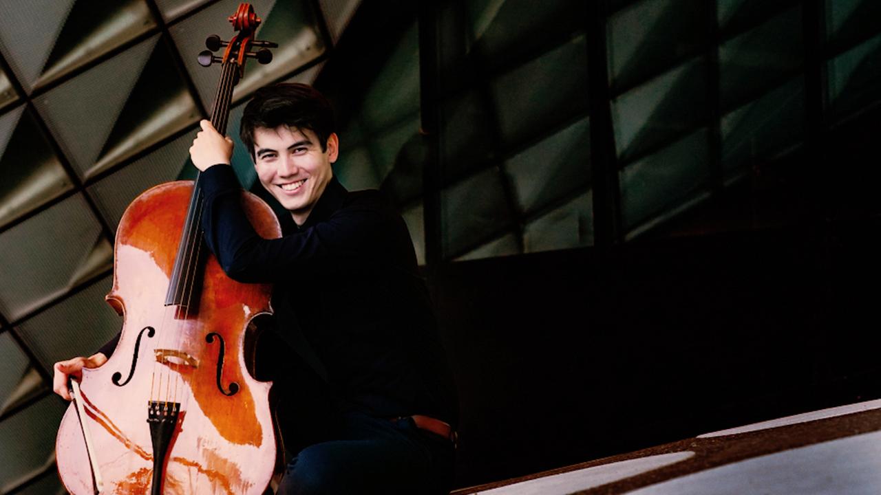 Ein junger Mann mit kurzen schwarzen Haaren lacht. Er hält ein Cello in der linken Hand. Hinter ihm erkennt man einen gemusterten Hintergrund. Ein junger Mann mit kurzen schwarzen Haaren lacht. Er hält ein Cello in der linken Hand. Hinter ihm erkennt man einen gemusterten Hintergrund.