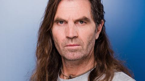 Der Profibergsteiger und Extremkletterer Thomas Huber hat lange Haare und einen Dreitagebart und blickt auffordernd in die Kamera.