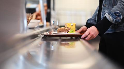 Eine Person steht in einer Kantine mit einem Tablett an der Essensausgabe. 