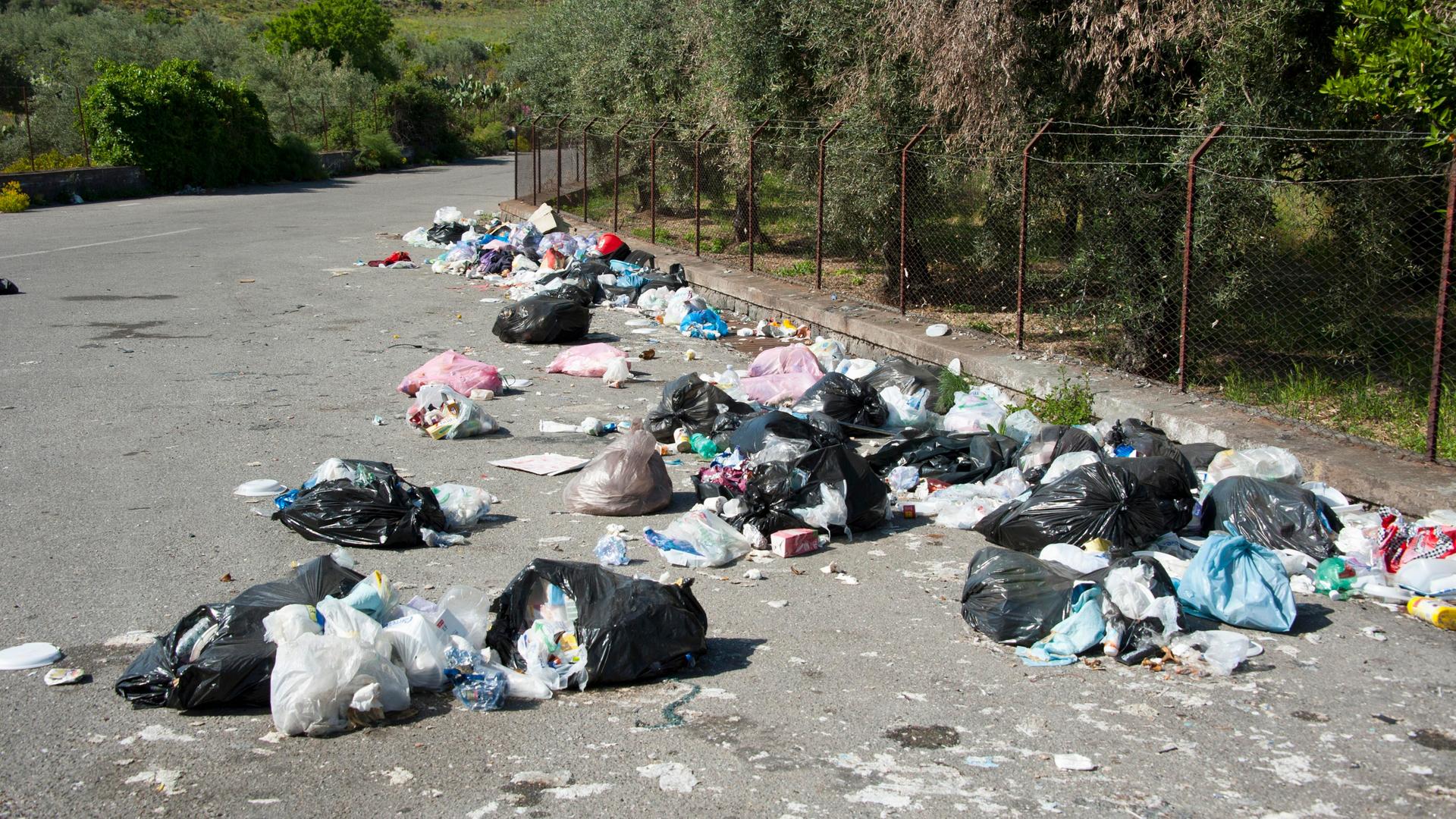 Am Rand einer Straße liegen viele Müll-Beutel. Die Beutel sind aufgerissen und der Müll liegt daneben. Die Straße ist umgeben von Bäumen. Es ist eine Straße auf der italienischen Insel Sizilien. Am Rand einer Straße liegen viele Müll-Beutel. Die Beutel sind aufgerissen und der Müll liegt daneben. Die Straße ist umgeben von Bäumen. Es ist eine Straße auf der italienischen Insel Sizilien.