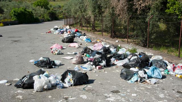 Am Rand einer Straße liegen viele Müll-Beutel. Die Beutel sind aufgerissen und der Müll liegt daneben. Die Straße ist umgeben von Bäumen. Es ist eine Straße auf der italienischen Insel Sizilien. 