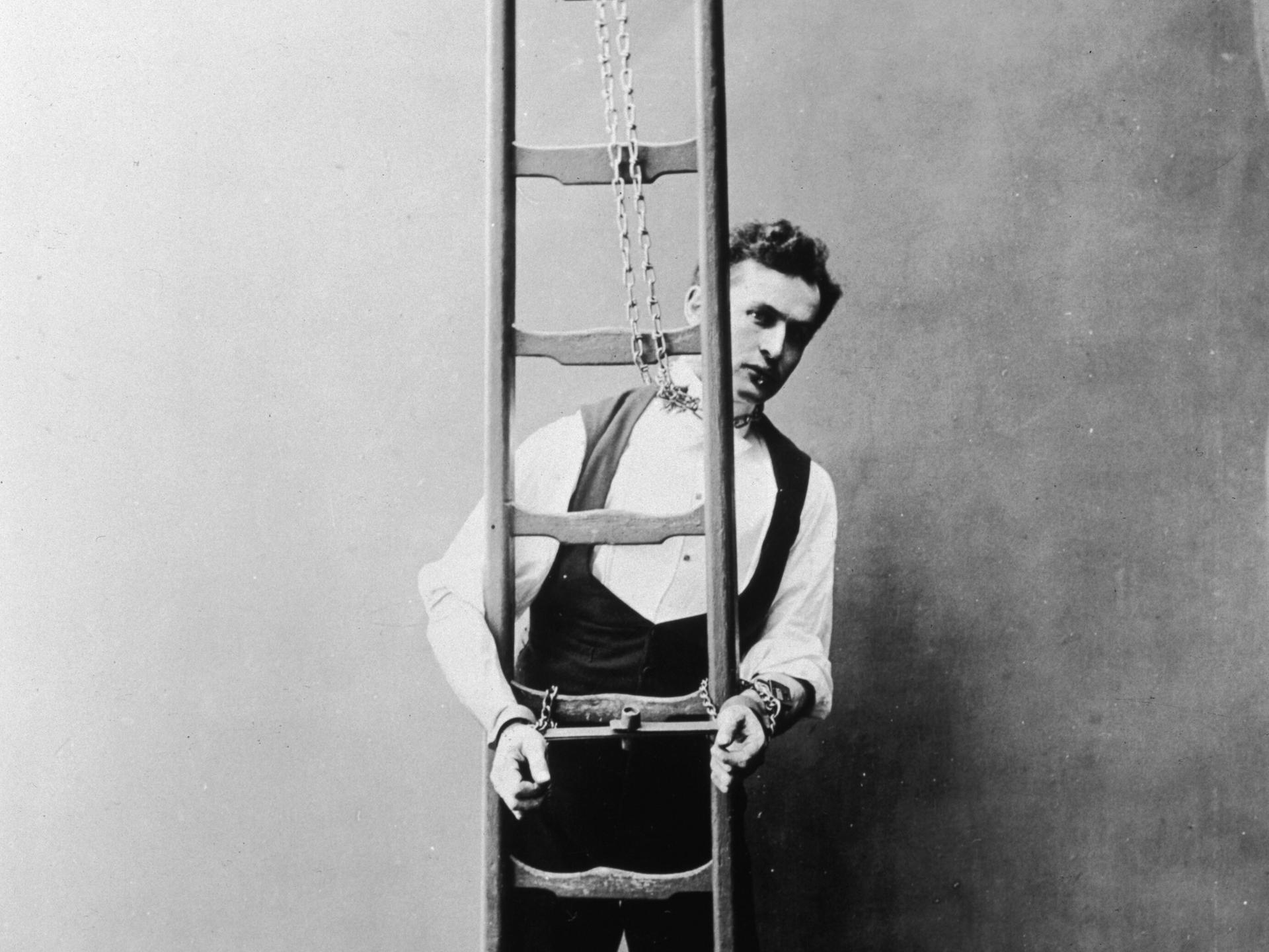 Historisches Schwarz-Weiß-Foto von Harry Houdini, der mit Ketten an Händen und Hals an eine Leiter gekettet ist.