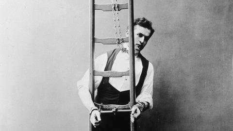 Historisches Schwarz-Weiß-Foto von Harry Houdini, der mit Ketten an Händen und Hals an eine Leiter gekettet ist.
