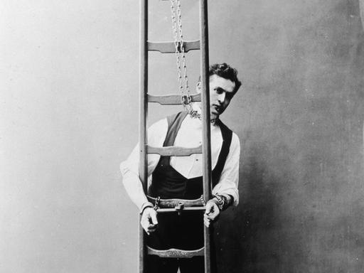 Historisches Schwarz-Weiß-Foto von Harry Houdini, der mit Ketten an Händen und Hals an eine Leiter gekettet ist.