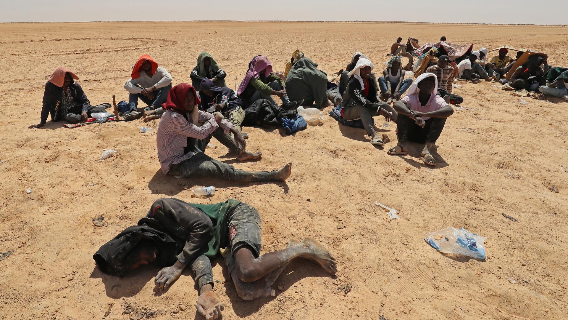 Menschen liegen oder sitzen im Wüstensand. Sie haben kaum eine Möglichkeit, sich vor der Hitze zu schützen.