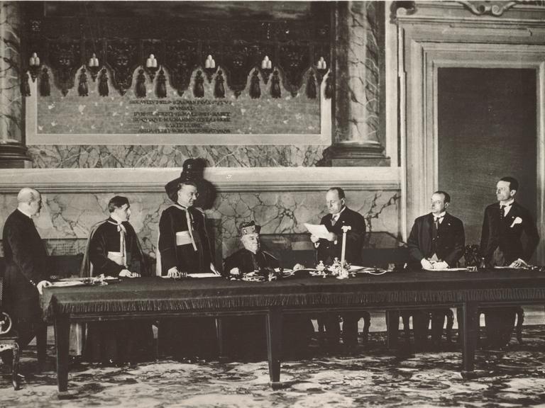 Vertragsunterzeichnung im Lateranspalast am 11. Februar 1929 mit Kardinalstaatssekretär Pietro Gasparri und dem faschistischen Ministerpräsidenten Italiens Benito Mussolini am Mikrofon stehend.