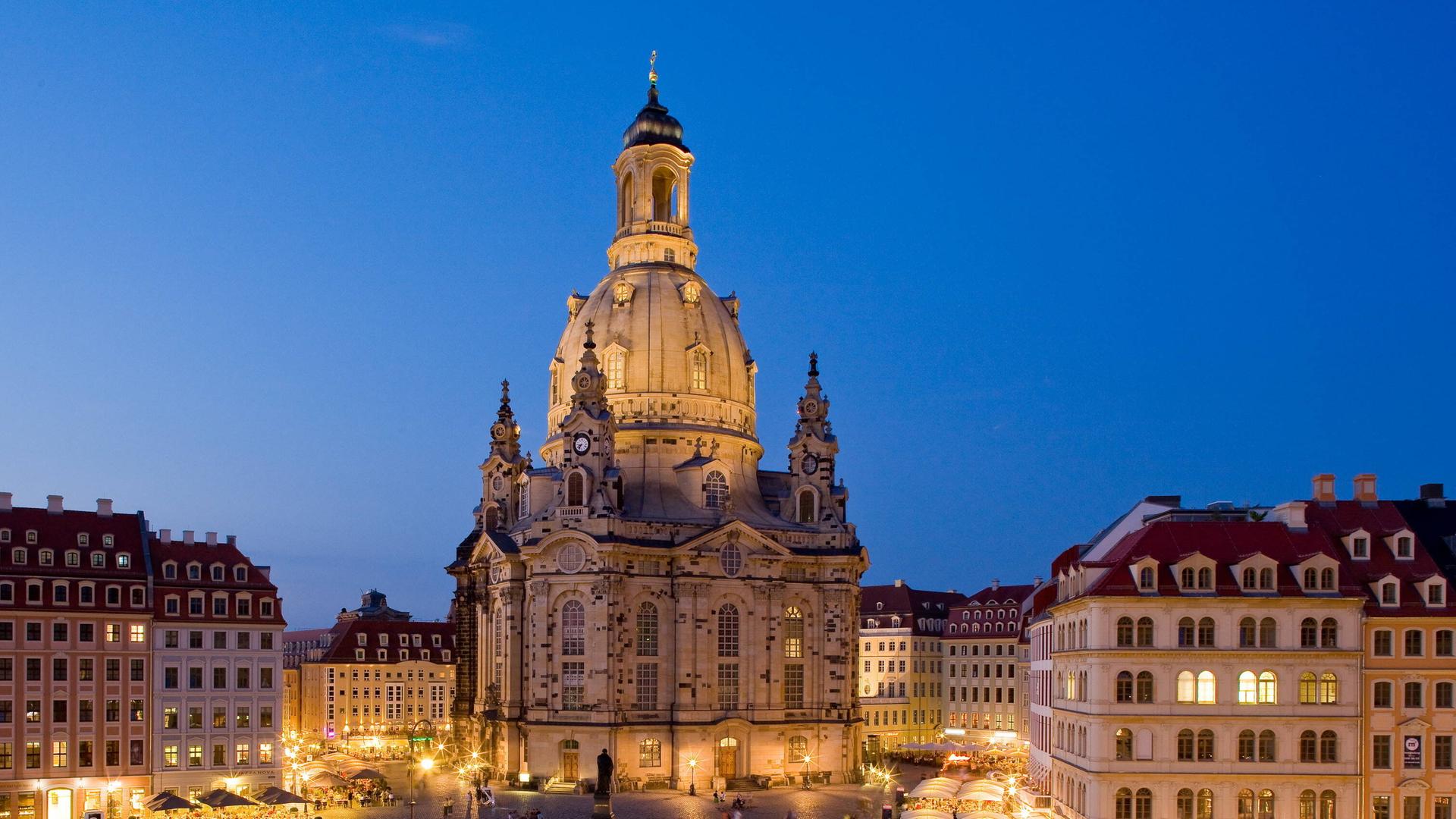 Blick auf den abendlichen, aber erleuchteten Neumarkt in Dresden auf die Frauenkirche vor blauem Himmel. 