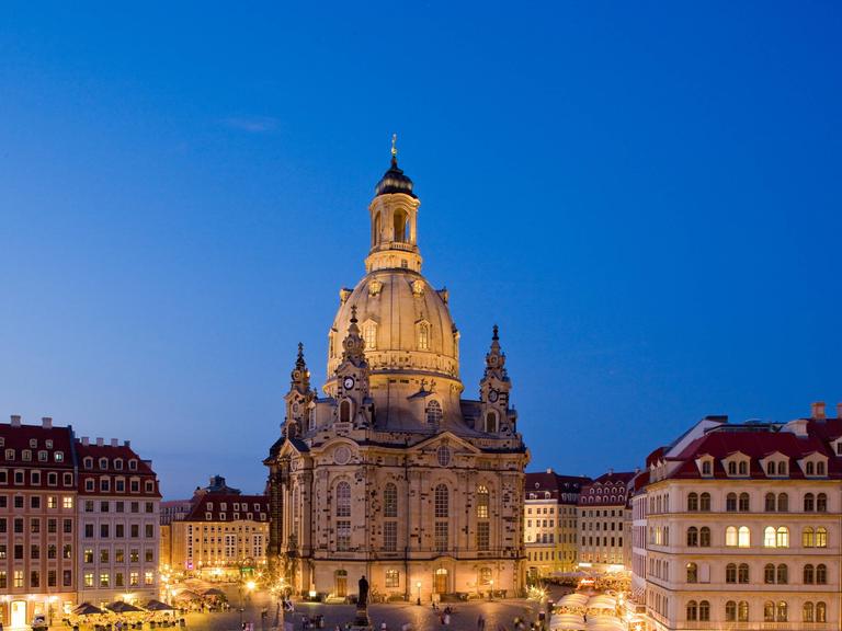 Blick auf den abendlichen, aber erleuchteten Neumarkt in Dresden auf die Frauenkirche vor blauem Himmel. 