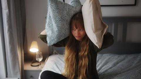 Eine Frau sitzt geqält im Bett und hält sich an jedes Ohr ein Kissen.