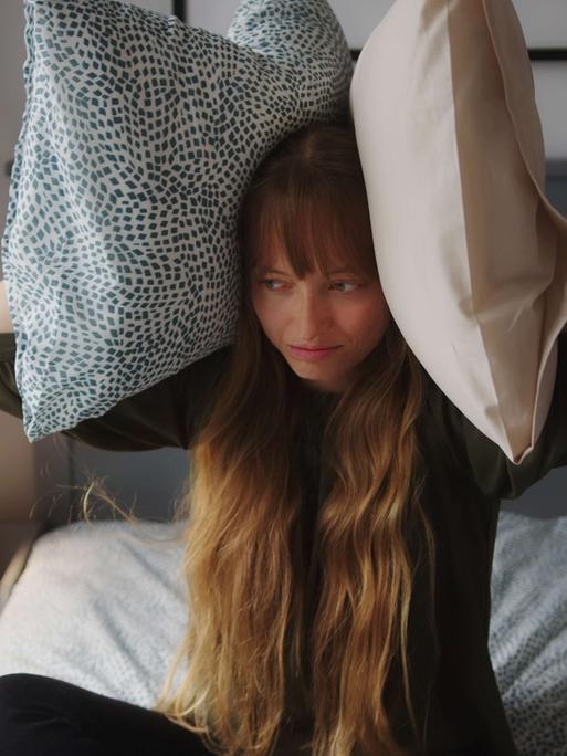 Eine Frau sitzt geqält im Bett und hält sich an jedes Ohr ein Kissen.