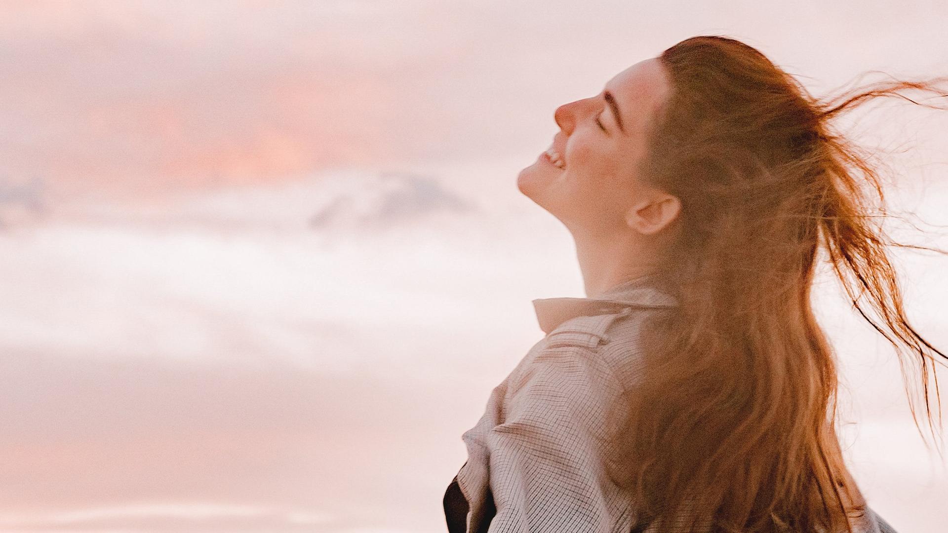 Eine junge Frau mit langen Haaren atmet glücklich die Luft des Abends mit rosa Wolken ein. 