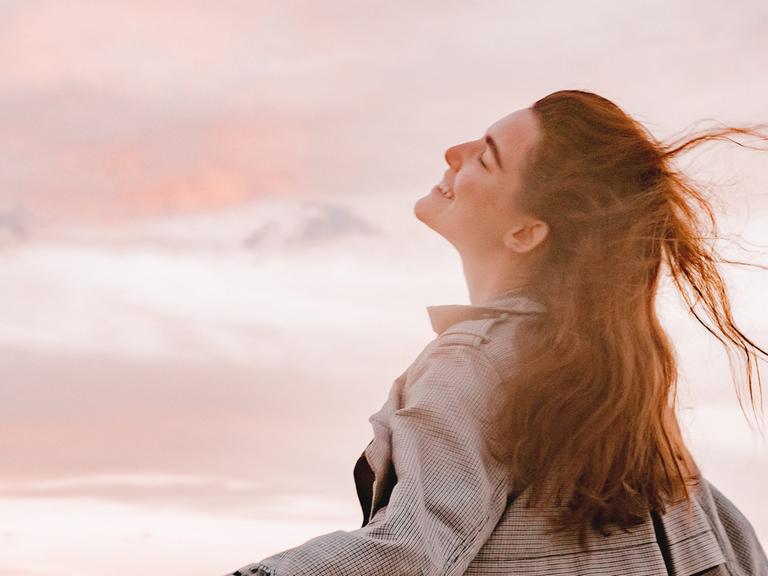 Eine junge Frau mit langen Haaren atmet glücklich die Luft des Abends mit rosa Wolken ein. 
