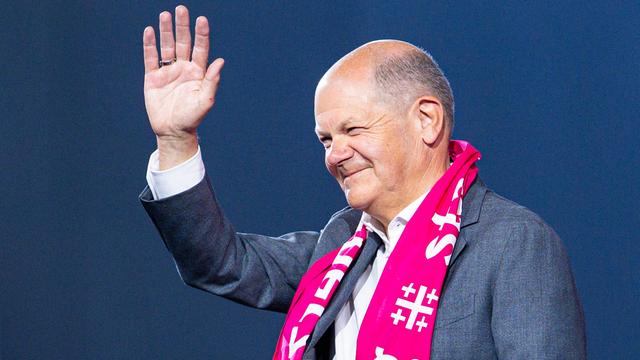 Ein lächelnder Bundeskanzler Olaf Scholz trägt einen pinken Schal mit dem Motto des Evangelischen Kirchentags und winkt.