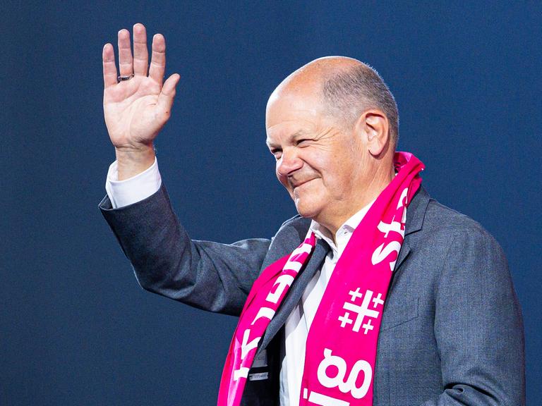 Ein lächelnder Bundeskanzler Olaf Scholz trägt einen pinken Schal mit dem Motto des Evangelischen Kirchentags und winkt.