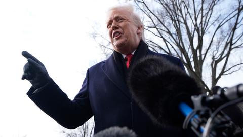 Das Bild zeigt Donald Trump aus der Froschperspektive. Der US-Präsident trägt einen dunklen Mantel und Handschuhe und hebt entschieden den Zeigefinger. Im Hintergrund: ein Baum ohne Blätter sowie der Himmel.