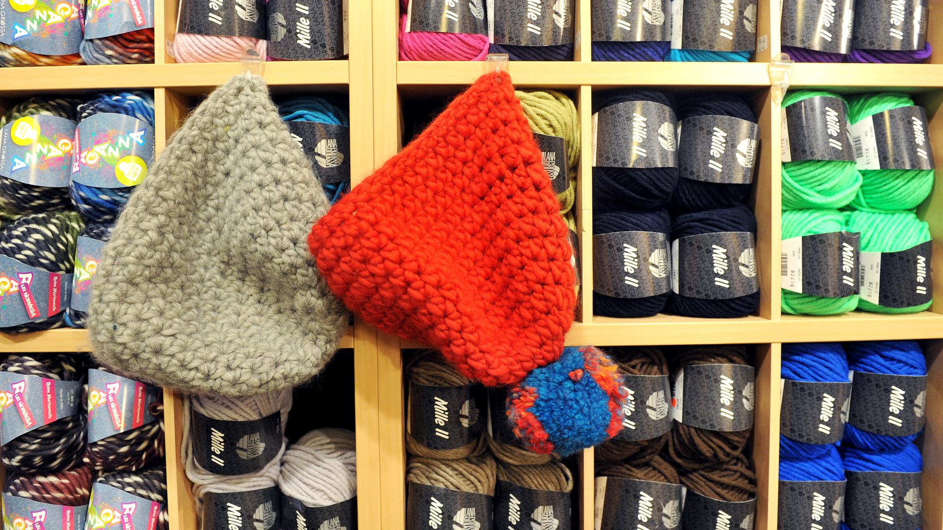 Zwei Wollmützen, eine graue und eine rote mit Bommel, hängen in einem Wollladen neben bunten Wollknäueln. Zwei Wollmützen, eine graue und eine rote mit Bommel, hängen in einem Wollladen neben bunten Wollknäueln.