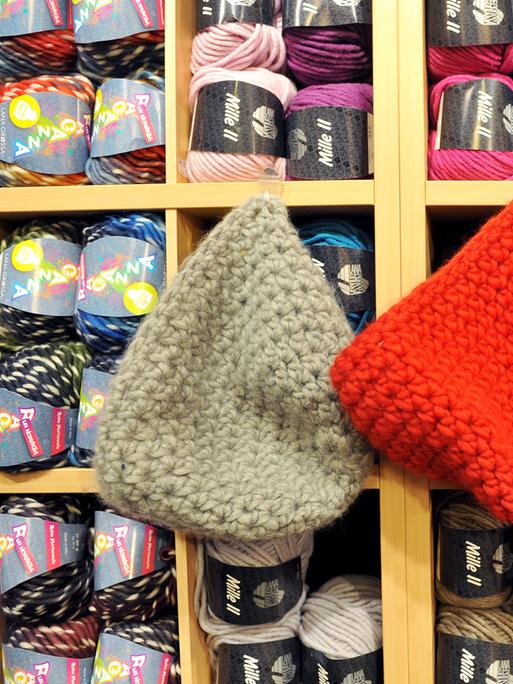 Zwei Wollmützen, eine graue und eine rote mit Bommel, hängen in einem Wollladen neben bunten Wollknäueln.