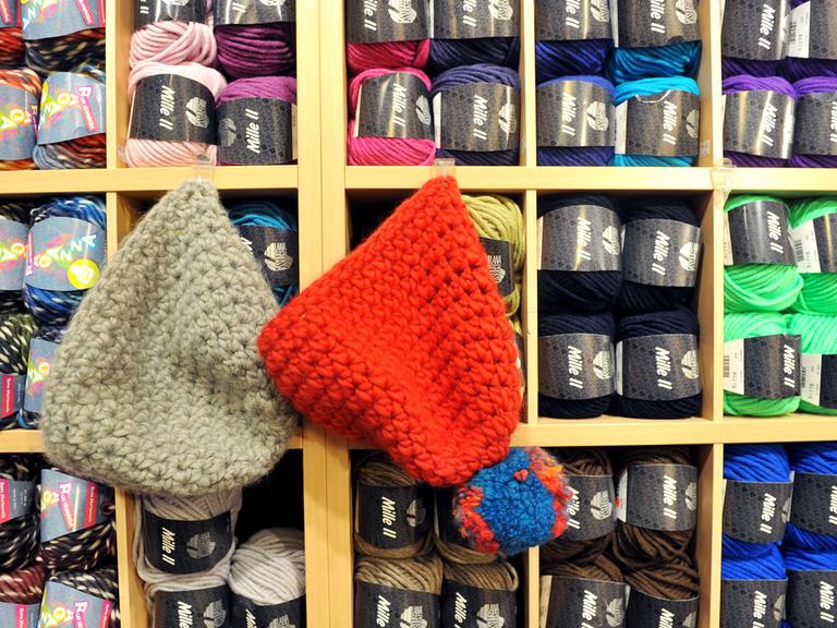 Zwei Wollmützen, eine graue und eine rote mit Bommel, hängen in einem Wollladen neben bunten Wollknäueln.