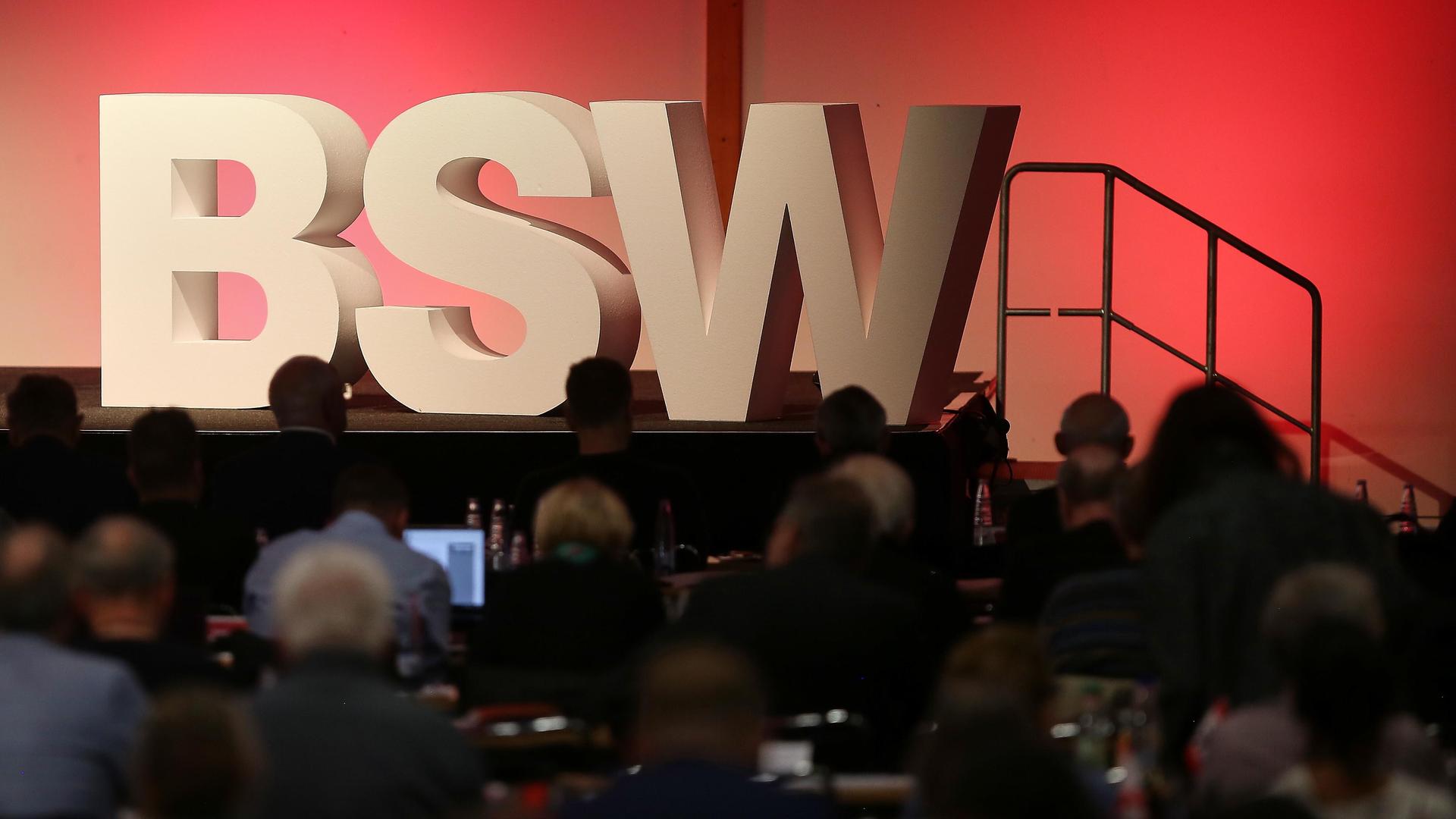 Das Logo des BSW in Großbuchstaben-Aufstellern steht auf einer Bühne. Im Vordergrund sitzen mehrere Delegierter im Schatten der Scheinwerfer.