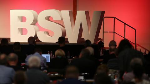 Das Logo des BSW in Großbuchstaben-Aufstellern steht auf einer Bühne. Im Vordergrund sitzen mehrere Delegierter im Schatten der Scheinwerfer.