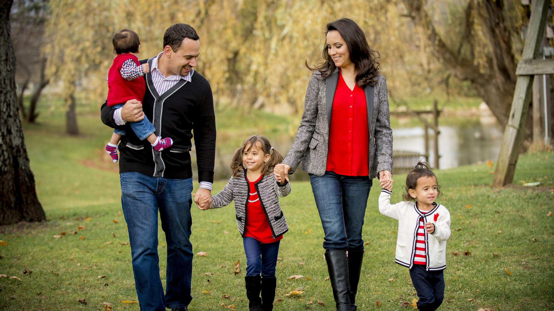 Eine Familie geht in einem Park spazieren, Vater, Mutter und drei Kinder sind lächelnd und händehaltend unterwegs.