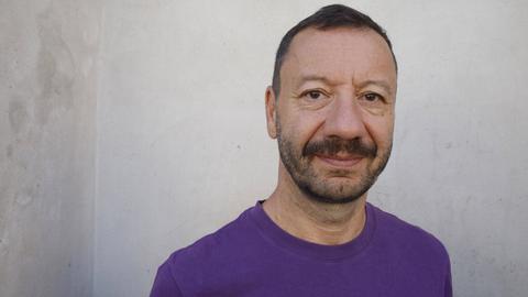 Portraitfoto von Selim Özdogan vor einer weiß-grauen Wand. er trägt einen lilafarbenen Pullover und schaut ernst in die Kamera.