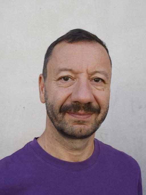 Portraitfoto von Selim Özdogan vor einer weiß-grauen Wand. er trägt einen lilafarbenen Pullover und schaut ernst in die Kamera.