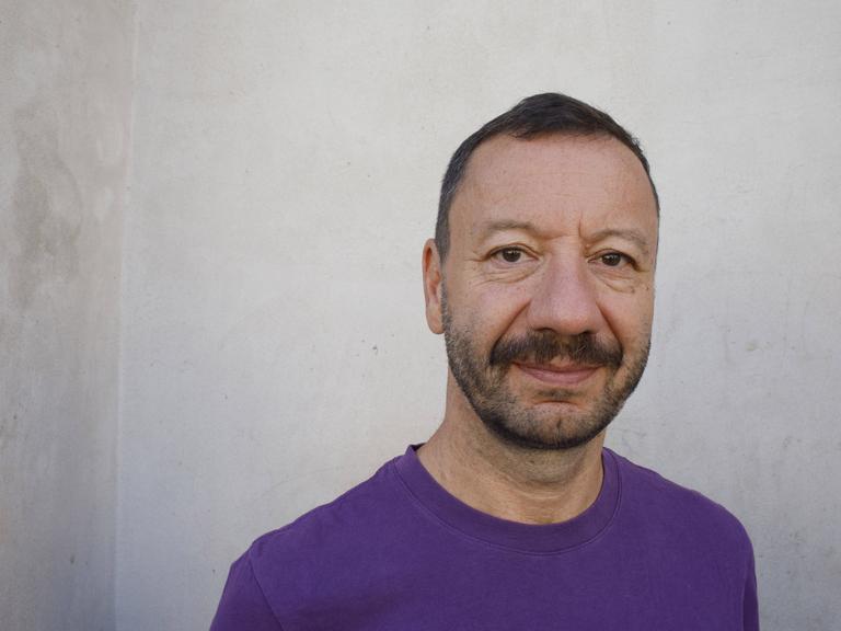 Portraitfoto von Selim Özdogan vor einer weiß-grauen Wand. er trägt einen lilafarbenen Pullover und schaut ernst in die Kamera.