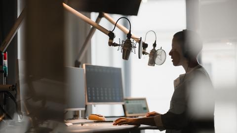 Eine junge Frau mit Kopfhörern steht in einem Aufnahmestudio am Schreibtisch und spricht etwas in Mikrofone ein